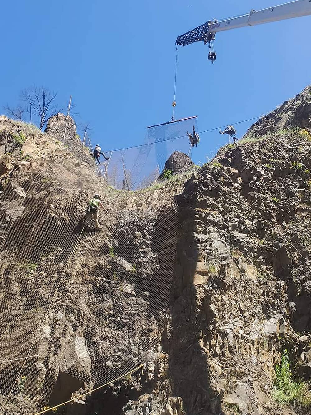 A photo of attenuator mesh being installed on hillside by workers and a crane