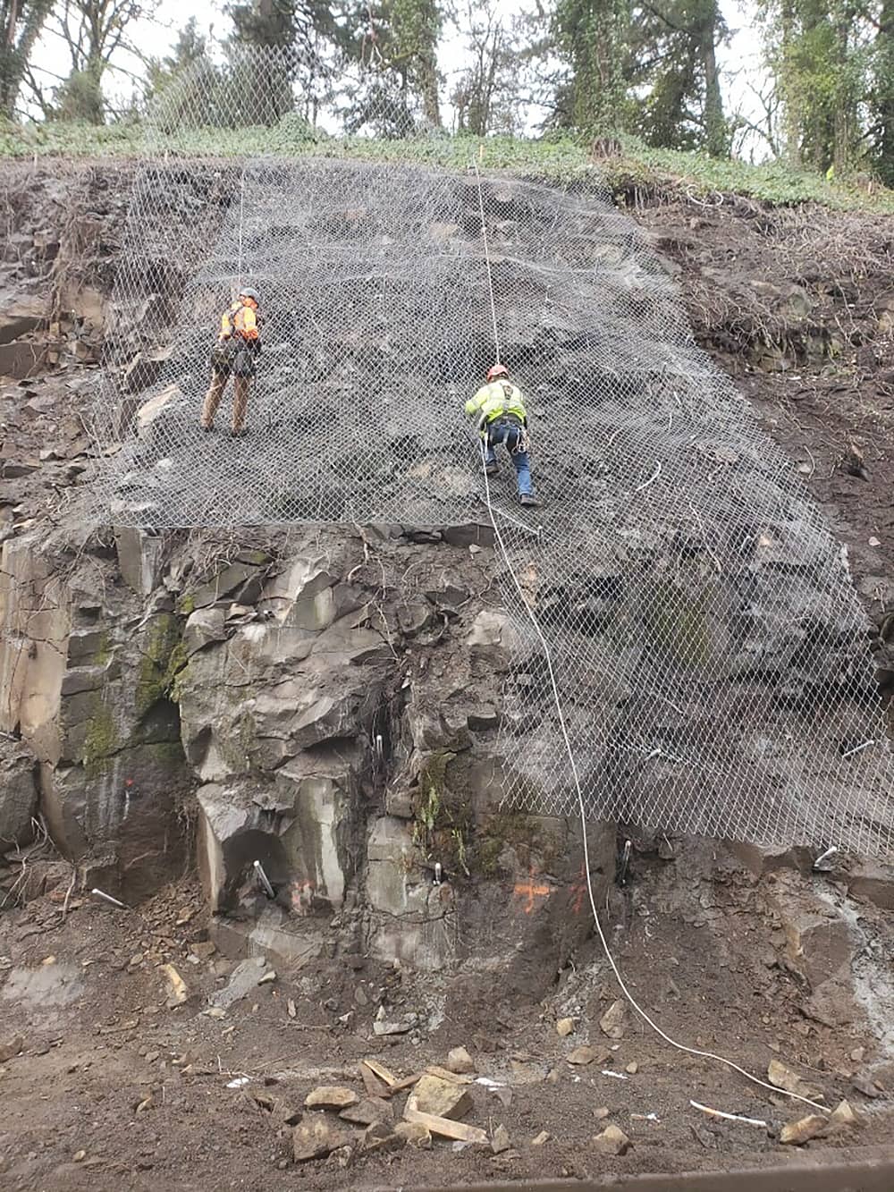 A photo of a trained workers installing draped mesh over a road cut next to a highway