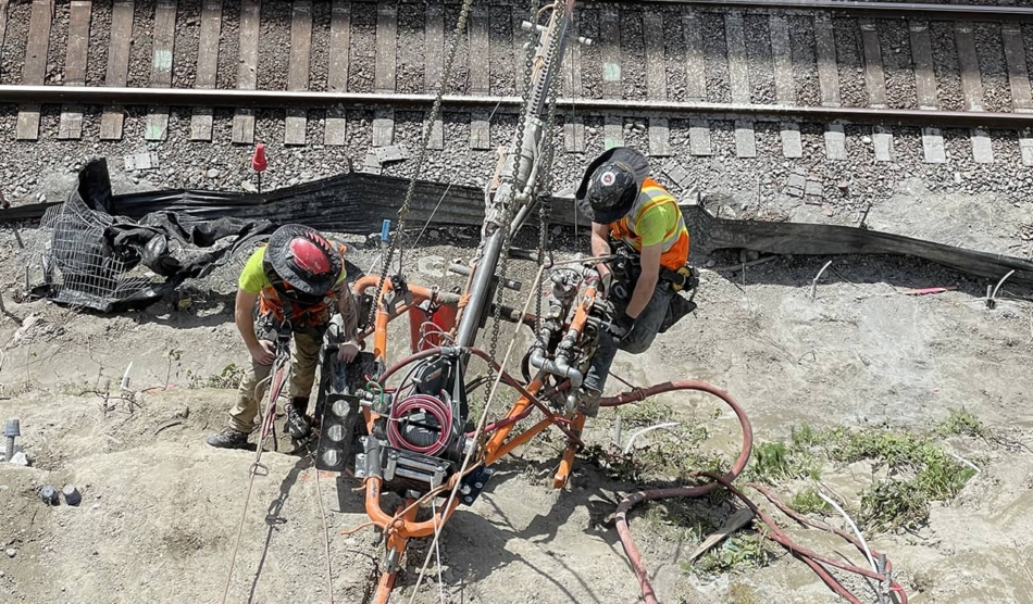 A photo of two men working near railroad tracks with a remote access drill