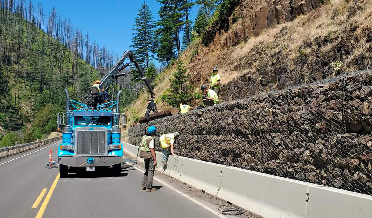 A photo of workers installing mesh, rock and log protective barrier along a highway