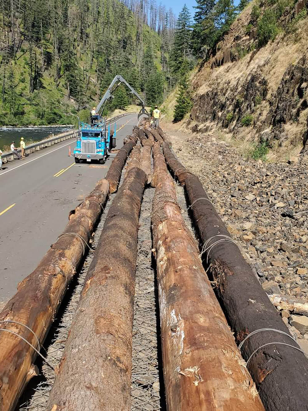 A photo of a truck with crane preparing to lift logs onto a roadside protective barrier