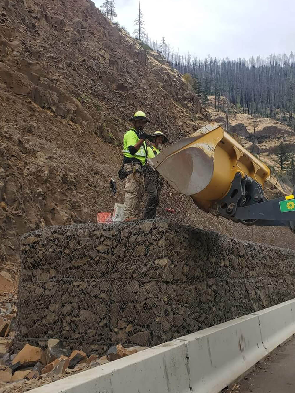 A photo of a workers installing a mesh and rock protective barrier along a highway