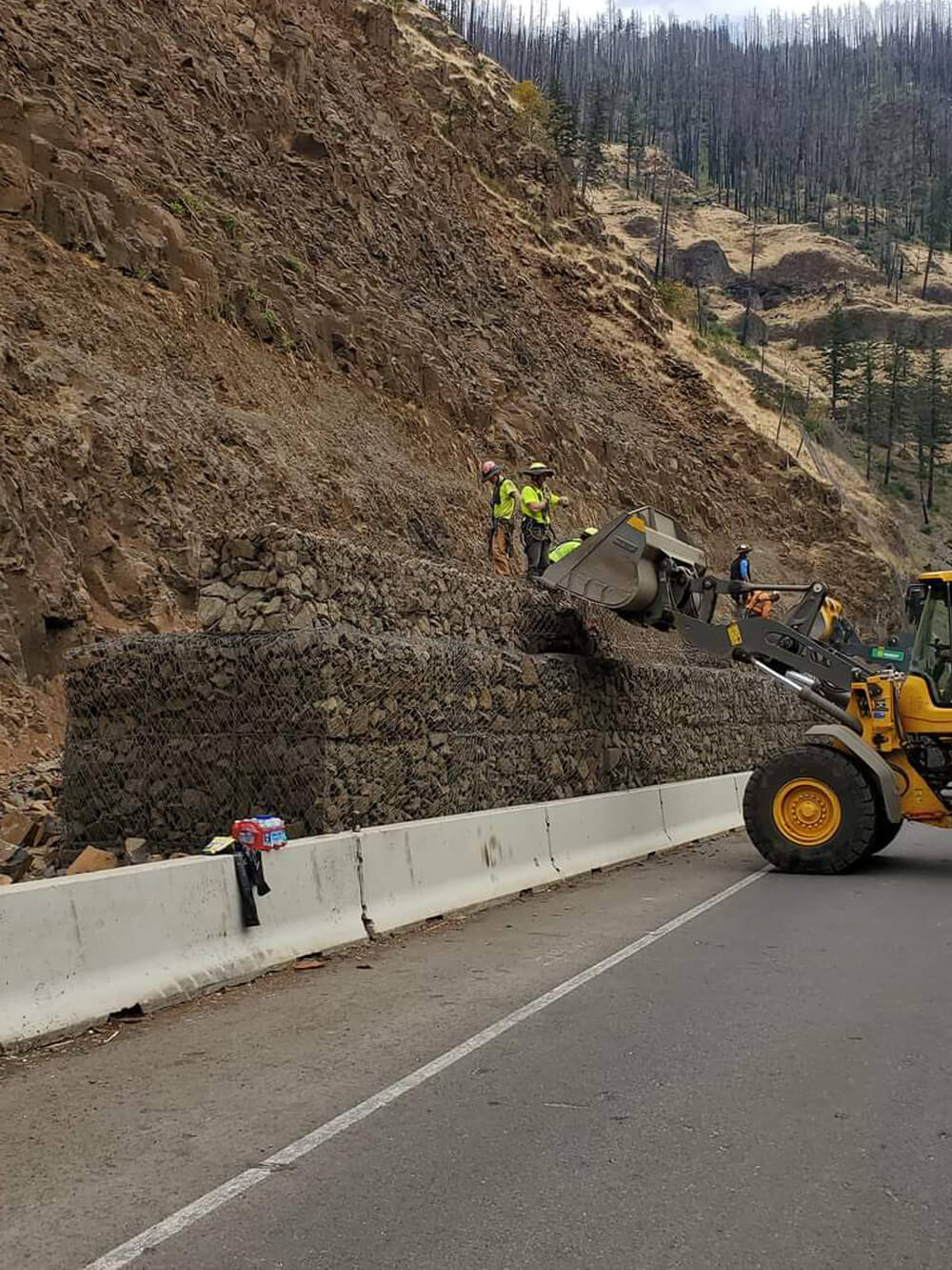 A photo of a workers and a bulldozer installing a mesh and rock protective barrier along a highway