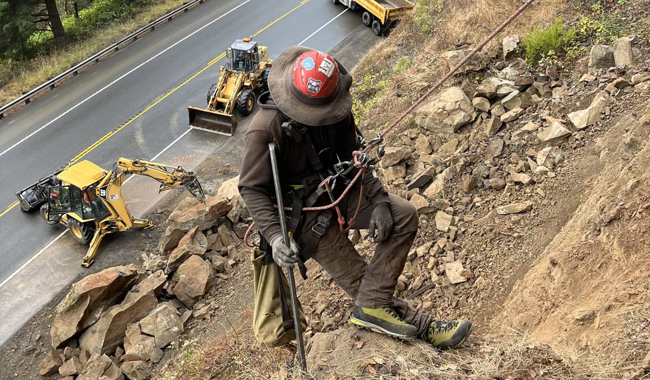 A photo of a trained professional performing rope access work on a road cut near a highway while bulldozers and other heavy equipment works to remove rock debris