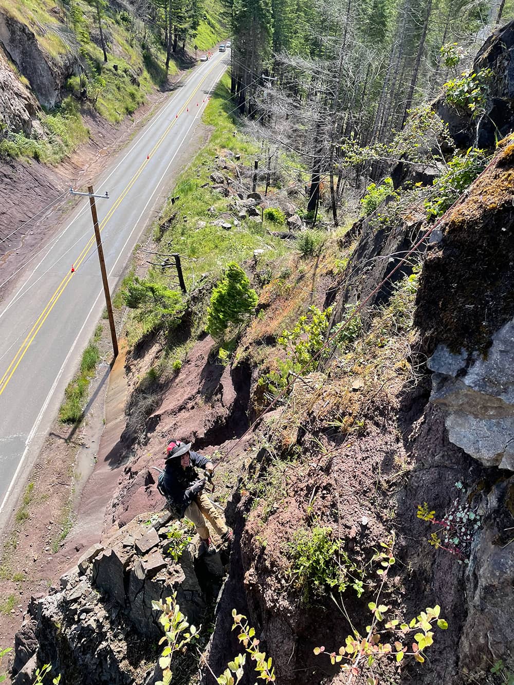 A photo of a trained professional performing rope access and rockfall mitigation work on a road cut near a highway